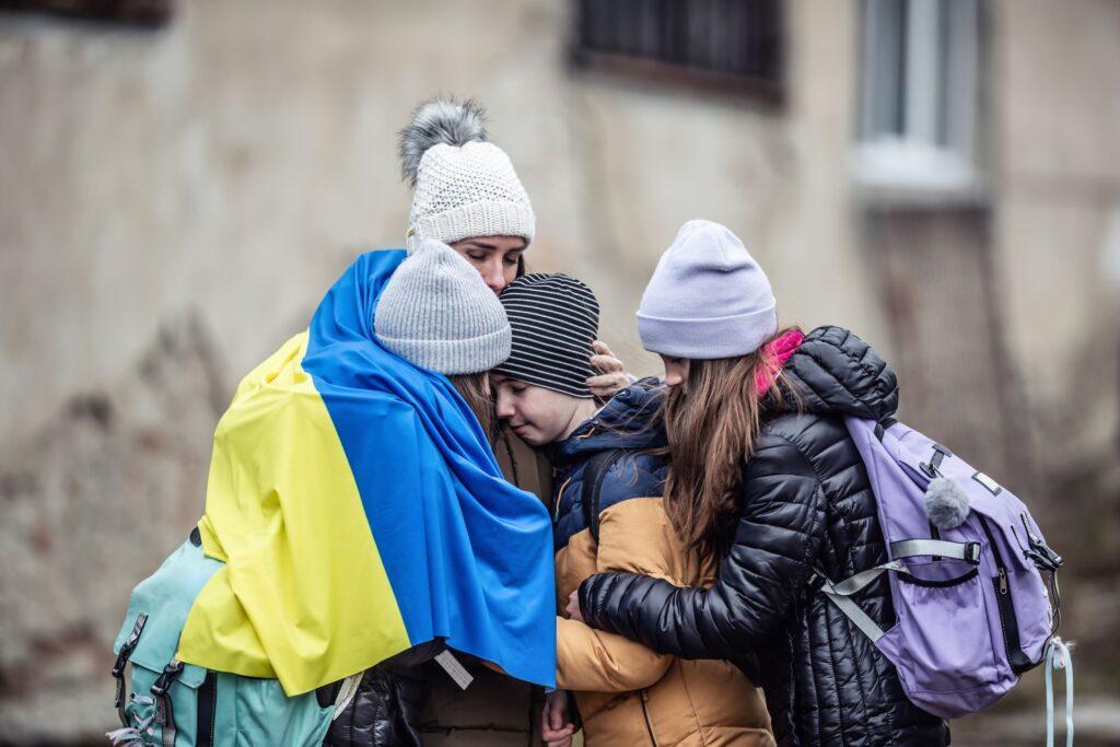 Eine geflüchtete ukrainische Mutter umarmt ihre Kinder, während sie in warme Kleidung gehüllt sind und eine ukrainische Flagge tragen. Der Ukrainekrieg hat schwere humanitäre Folgen und treibt viele Familien in die Flucht. Die EU-Sanktionen und die Finanz Sanktionsliste spielen eine entscheidende Rolle bei den wirtschaftlichen Maßnahmen gegen Russland.